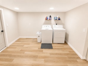 A laundry room with two washing machines, a dryer, a trash bin, and shelves with laundry supplies. - The First Inn Parker Arizona (Parker)