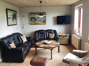 Ein Wohnzimmer mit einem schwarzen Ledersofa, einem braunen Ottomanen, einem Couchtisch mit Muster, einem schwarzen Ledersessel, einem Fernseher und einem Bild an der Wand.