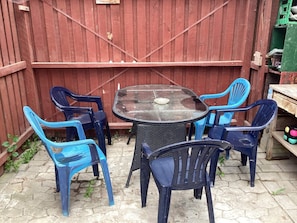Un patio con una mesa redonda y seis sillas, una cerca de madera y un cesto verde al fondo.