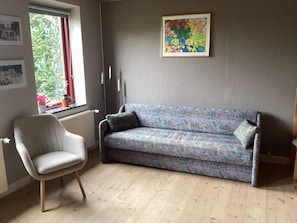 Ein Zimmer mit einer gemusterten Couch, einem weißen Sessel, einem Fenster mit Blick auf Grünflächen und einem gerahmten Kunstwerk an der Wand.