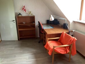 A room with a sloped ceiling, a wooden desk with a computer and lamp, a chair with a blanket, and a flag on the wall. - 6 Person Holiday Home in Bandholm (Bandholm)