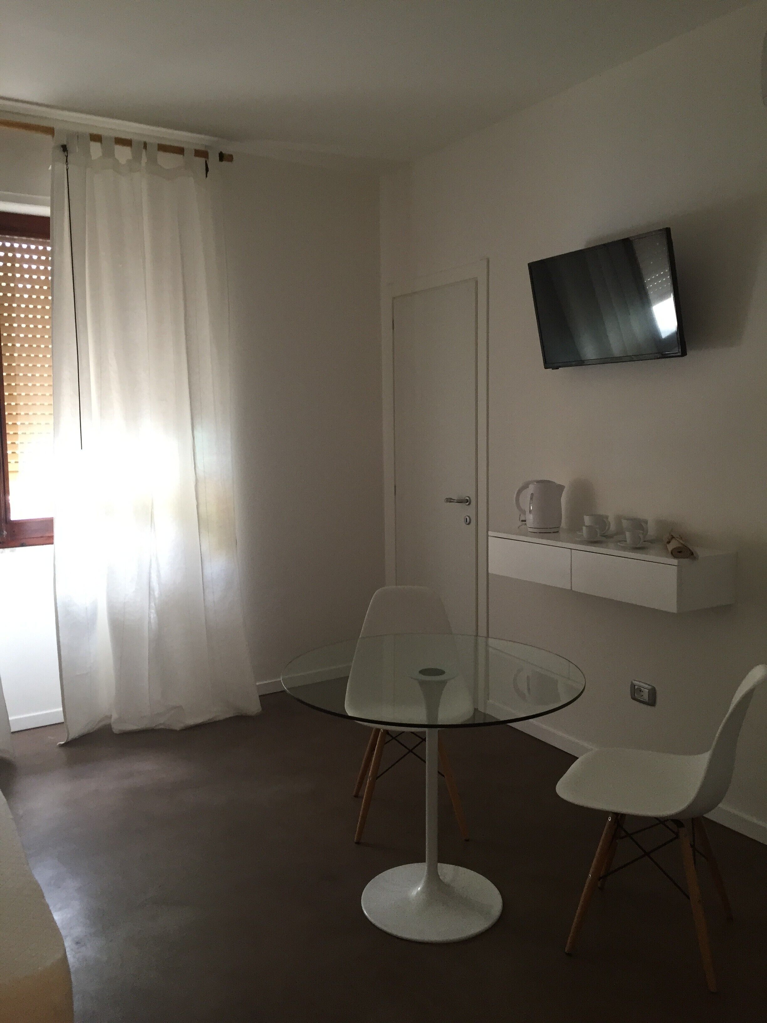 Een kamer met een glazen tafel, twee stoelen, een televisie aan de muur, een deur en een plank met bekers en een waterkoker.