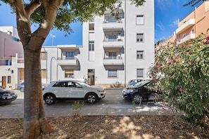 En moderne lejlighedsbygning med flere balkoner, parkerede biler og et træ i forgrunden.