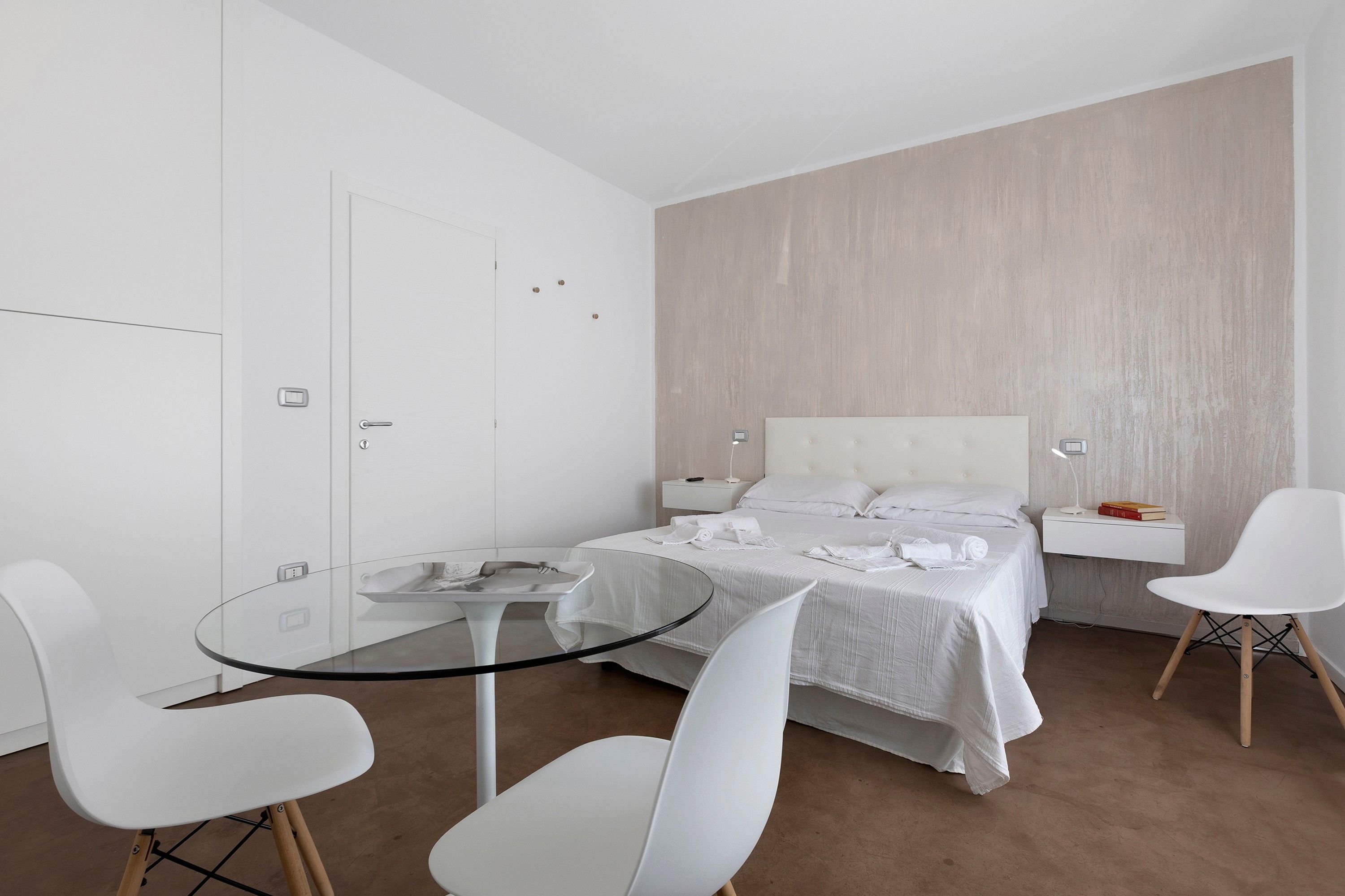 A modern hotel room with a bed, a glass table, two white chairs, a bedside table with a lamp, and a wall-mounted TV.