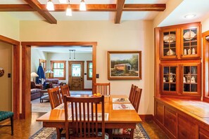 A dining area with a wooden table, chairs, and a rug. There is a kitchen area with wooden cabinets and a painting on the wall.