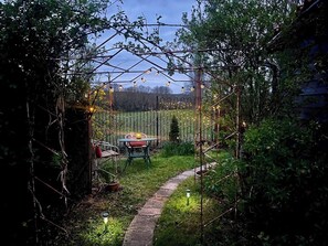 Um jardim com um caminho, uma mesa com cadeiras e luzes de cordão.