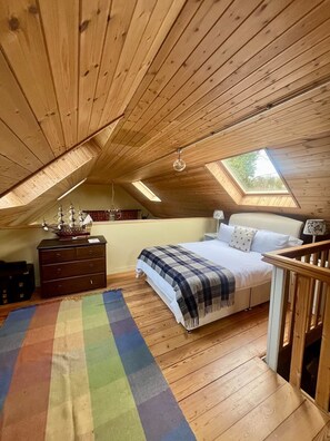Ein Schlafzimmer mit geneigter Decke, einem Bett, einer Kommode, einem bunten Teppich und einem Oberlicht.