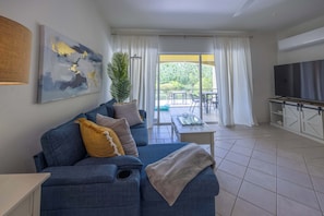 A modern living room with a sofa, a coffee table, and a TV. There is a sliding glass door leading to an outdoor area with a table and chairs. - Grace Bay Condo with Pool and Patio Near Beach (Grace Bay)