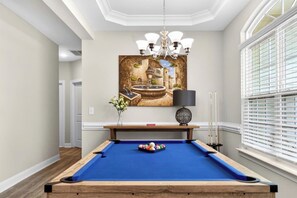A pool table with a blue felt surface, a chandelier, and a painting of a fountain on the wall.