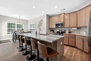 A modern kitchen with a central island, bar stools, wooden cabinets, stainless steel appliances, and a large window.