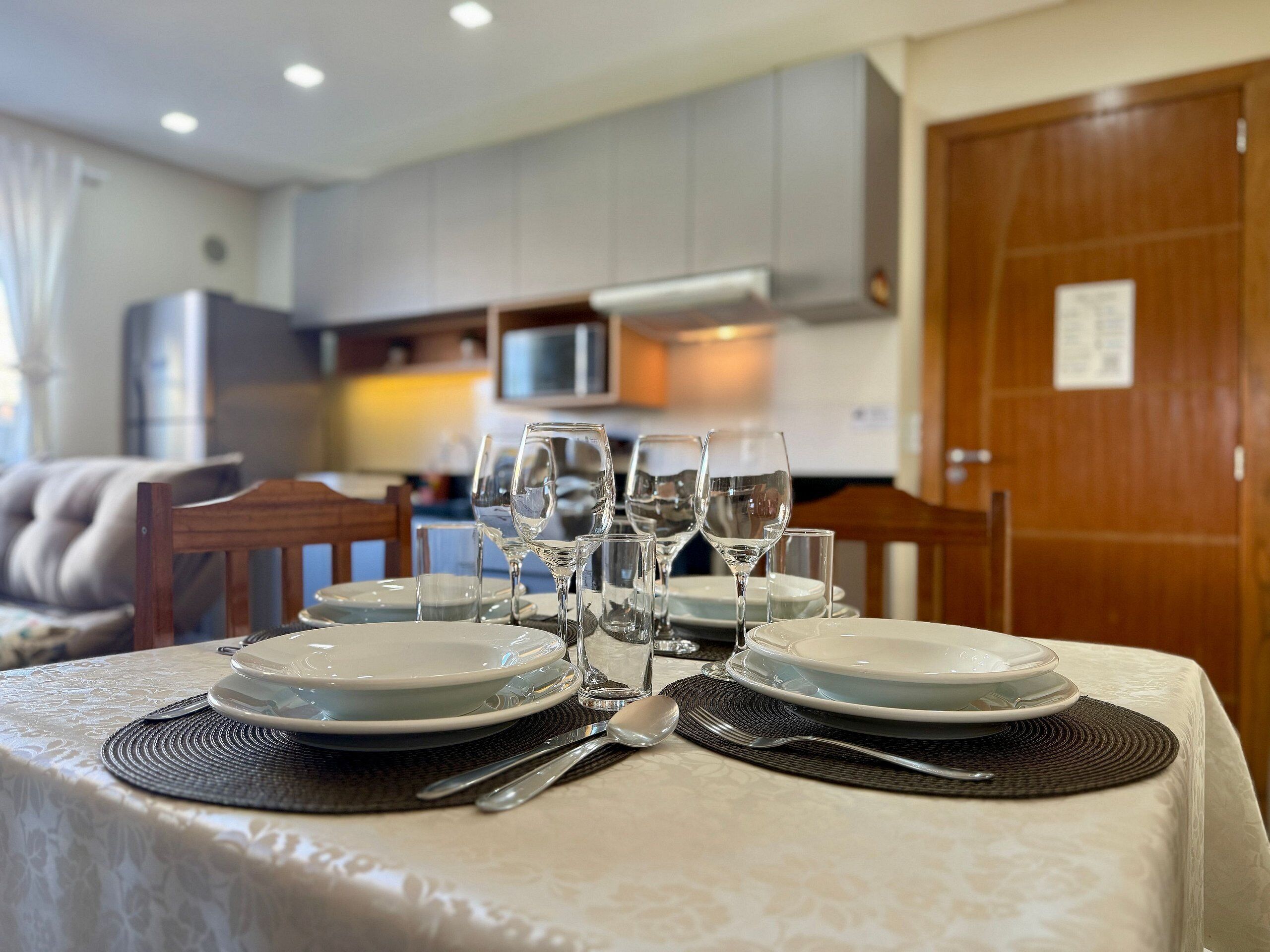 Uma mesa de jantar posta com pratos, copos e talheres, com uma cozinha ao fundo.
