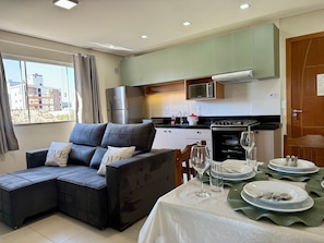Una sala moderna con un sofá seccional, una mesa de comedor para dos personas y una zona de cocina con microondas, estufa y refrigerador.