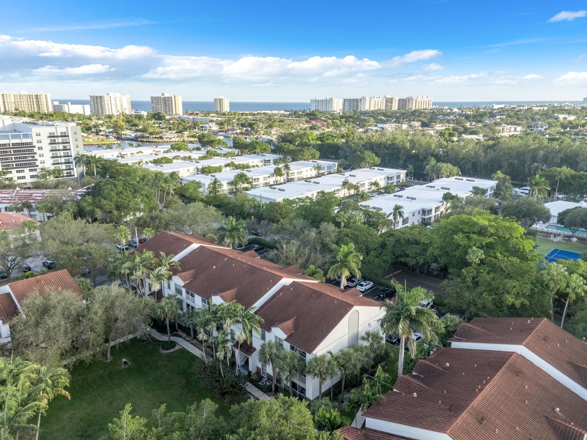 A residential area with multiple buildings, including a large white building with a red roof, surrounded by greenery and palm trees.