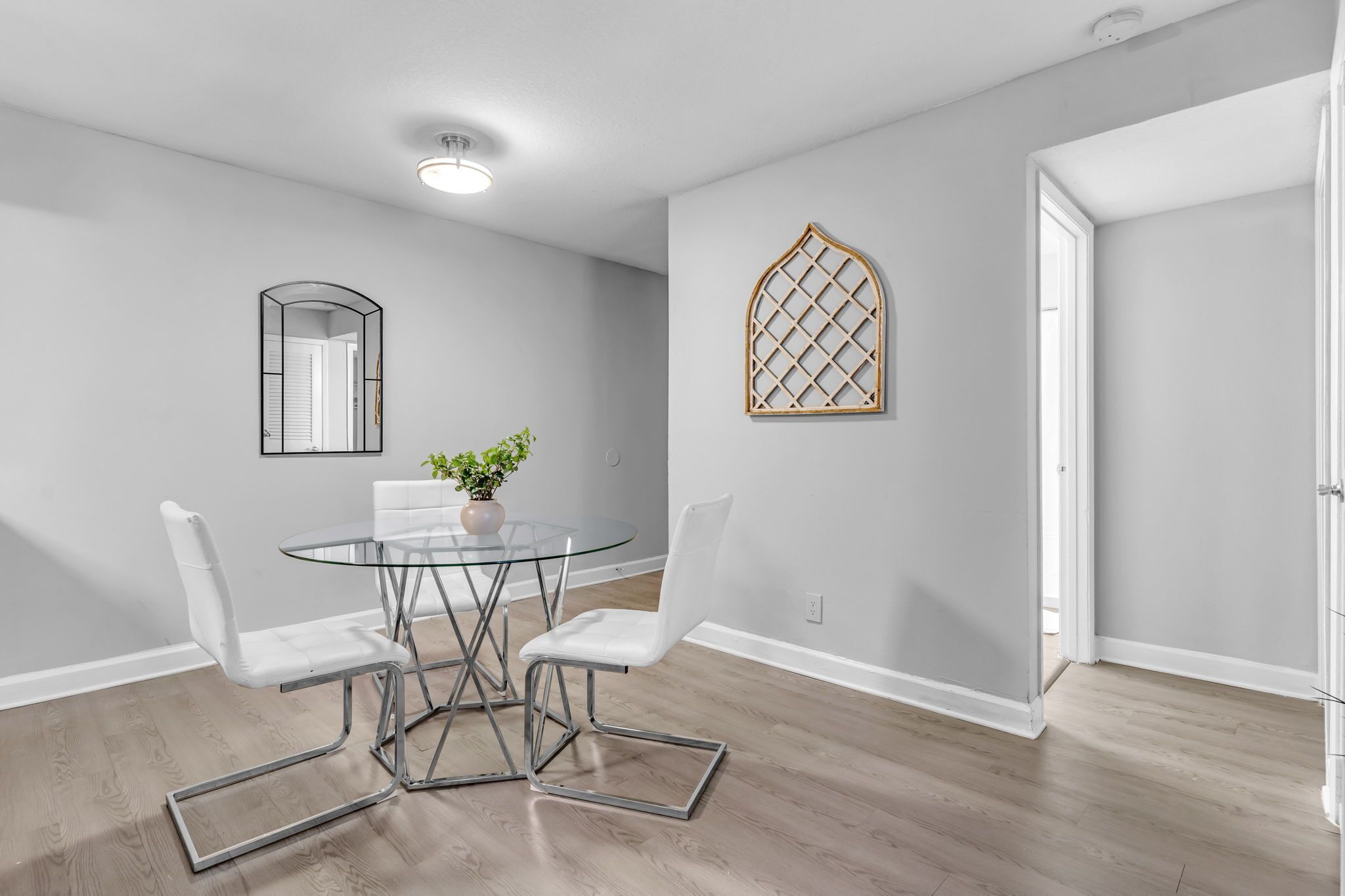 A dining area with a glass table and white chairs, a mirror, and a decorative wooden lattice on the wall.