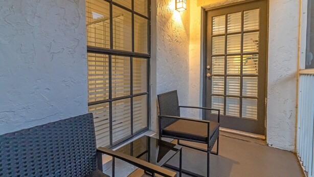 Een balkon met een glazen tafel en stoelen, een deur met een raam en een wandlamp.