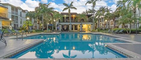A swimming pool with lounge chairs, palm trees, and a building with balconies in the background.