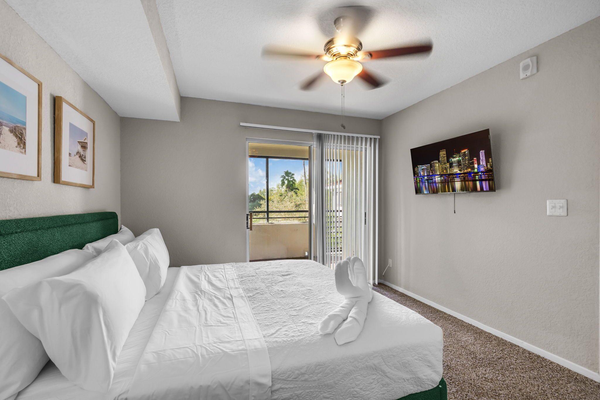 A hotel room with a bed, a ceiling fan, a TV, and a balcony with a view of the city.