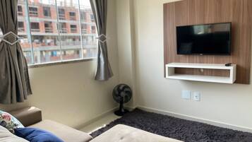 En stue med sofa, tv ophængt på væggen, et vindue med gardiner og en ventilator.