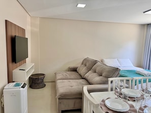 A compact hotel room with a flat-screen TV mounted on the wall, a sofa, a small dining table set with plates and glasses, and a bed with a green bedspread. - 103 | Res. 287 • Affordable for up to 7 People, Close to the Beach (Bombinhas)