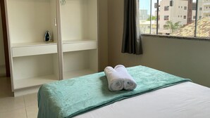 A hotel room with a bed, a green blanket, a pair of towels, a wardrobe, and a window with a view of buildings. - 287.203 - AP Aurora: Affordable, cozy and just steps from the beach (Bombinhas)