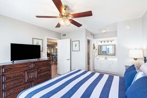 Ein Schlafzimmer mit Bett, Fernseher, einem hölzernen Schminktisch, Deckenventilator, einem Badezimmer mit Waschbecken und Spiegel sowie einem Essbereich im Hintergrund.