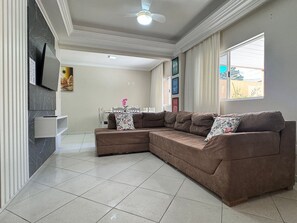 A living room with a sectional sofa, a flat-screen TV, a dining area with a table and chairs, and a large window with curtains. - 01 | Res. 364 • Ground floor - 3 bedrooms - 5min walk to the Sea (Bombinhas)