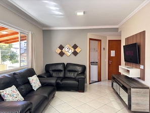A living room with a black leather sofa, a flat-screen TV mounted on a wooden wall, and a view of a residential area through a large window. - 03 | Apt. 364 • Apt. with Balcony - 2 Bedrooms - 5 min Walk to the Sea (Bombinhas)
