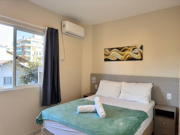 A hotel room with a bed, a painting on the wall, an air conditioning unit, and a window with curtains. - 03 | Apt. 364 • Apt. with Balcony - 2 Bedrooms - 5 min Walk to the Sea (Bombinhas)