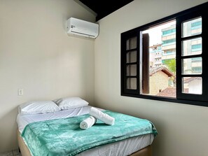 A single bed with white bedding and a teal blanket, an air conditioning unit on the wall, and a window with a view of buildings outside. - 09 | Res. 305 • Beautiful Apartment – Great Value, 5 Min from the Beach (Bombinhas)