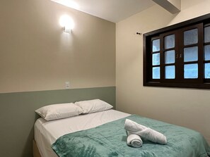 Ein Einzelbett mit grüner Bettdecke, ein aufgerolltes Handtuch, eine Wandleuchte und ein Fenster mit satinierten Scheiben.