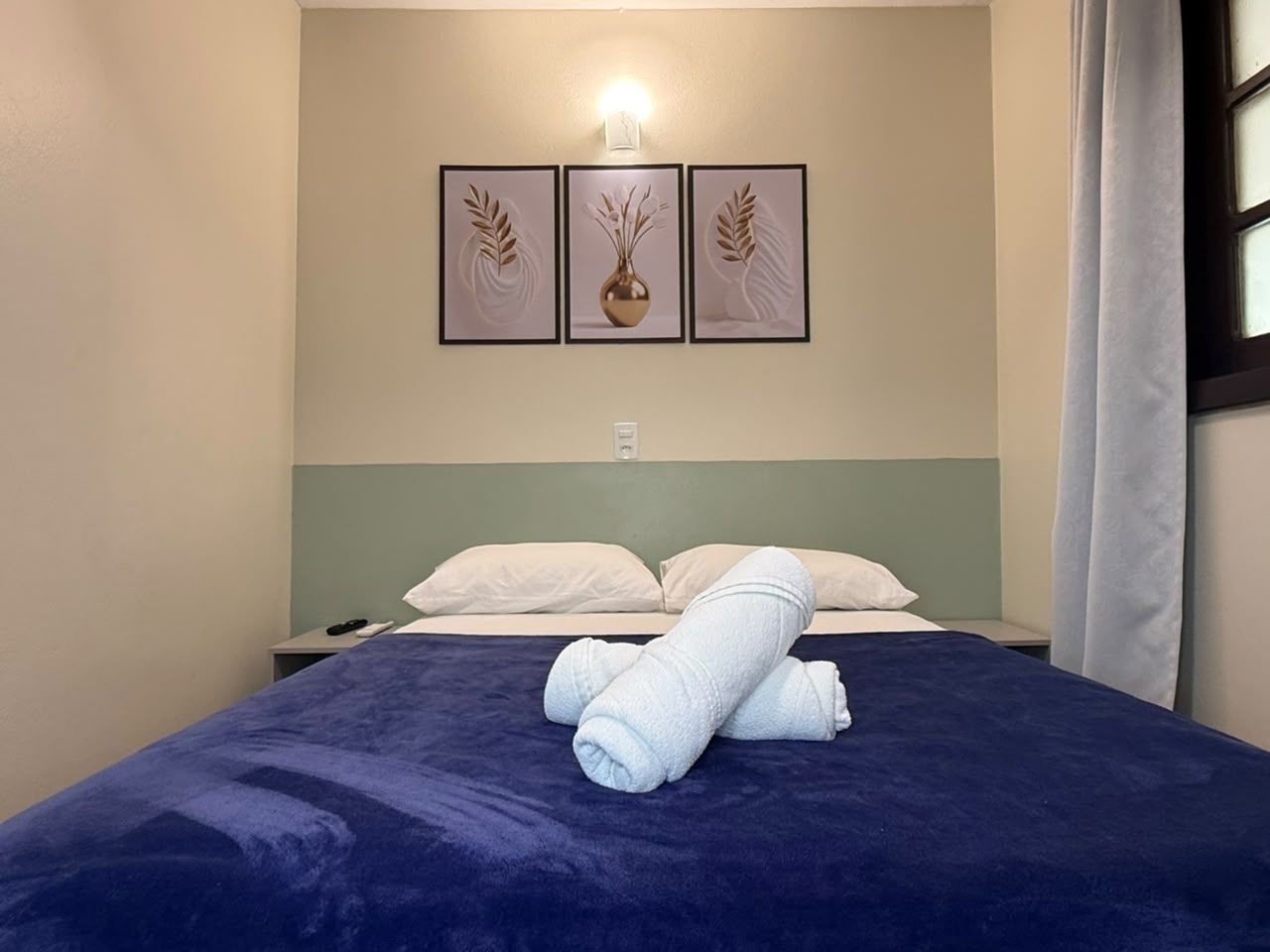 Quarto de hotel com cama, almofadas, mesa de cabeceira e luminária fixa na parede.