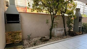 An outdoor patio area with a brick barbecue grill, a stone bench, and a small tree.