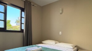 A single bed with a green blanket and two rolled towels, a nightstand with drawers, a window with curtains, and a wall-mounted light fixture. - 02 | Res. 305 • Cozy Apartment for Couple – 5min from the Beach (Bombinhas)