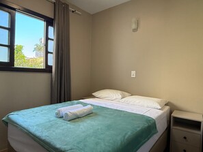 Ein Einzelbett mit grüner Decke und zwei gerollten Handtüchern, ein Nachttisch mit Schubladen, ein Fenster mit Vorhängen und eine wandmontierte Leuchte.