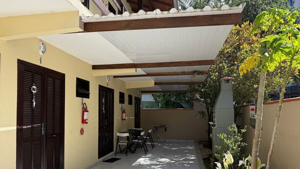 A covered patio with a tiled floor, a small table and chairs, a fire extinguisher, and a door with a security device. - 02 | Res. 305 • Cozy Apartment for Couple – 5min from the Beach (Bombinhas)