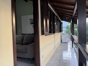 Ein überdachter Balkon mit einer Couch und einem Fernseher, Fliesenboden und Blick auf Grünflächen.