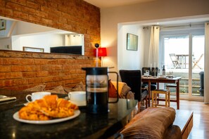 Ein gemütlicher Wohnbereich mit einer Ziegelwand, einem Couchtisch mit Croissants, einer Kaffeemaschine und einem Esstisch mit Stühlen.