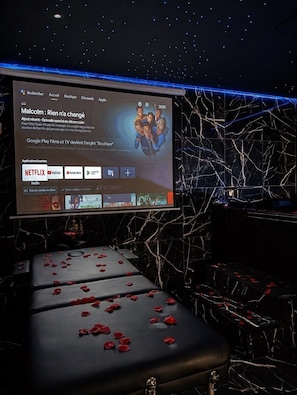 A hotel room with a large screen displaying a movie selection, a massage table with rose petals, and a dark, starry ceiling.