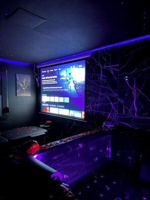A dark room with a large screen displaying a video game, a marble wall with a crack pattern, and a bar area with a drink dispenser. - Obsidian Suite/Air-Conditioned Love Room (Triel-sur-Seine)