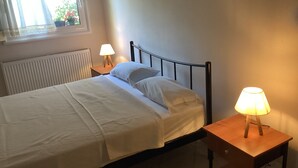 En säng för en person med ett vitt överkast, två sängbord med lampor, ett fönster med gardiner och ett element.
