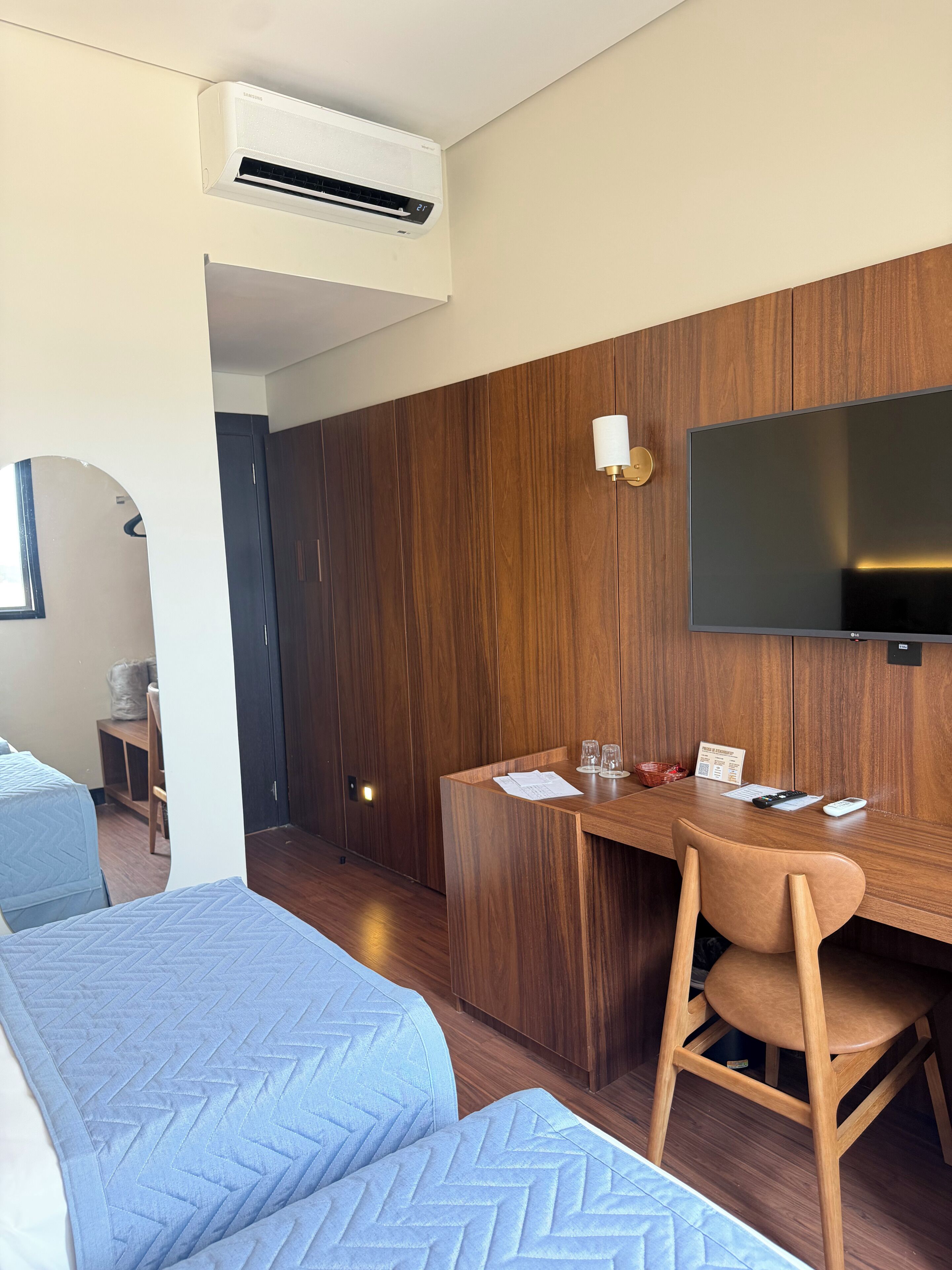 A hotel room with a bed, a desk, a chair, a television, and an air conditioning unit.
