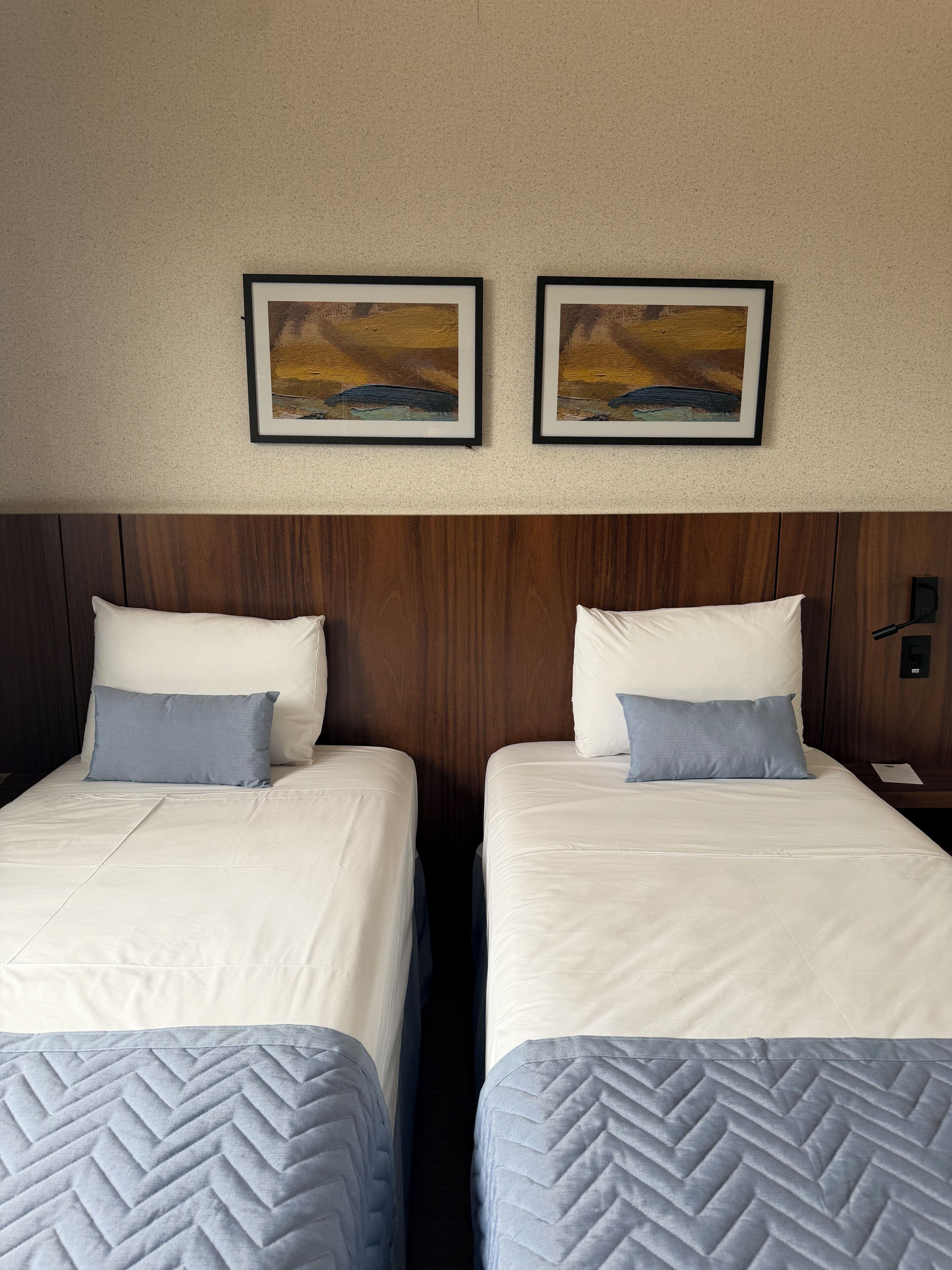 A hotel room with two beds, each with a pillow and a quilted bedspread. There are two framed paintings on the wall above the beds.