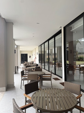 An outdoor seating area with tables and chairs, large windows, and a view of the outdoors. - Class Hotel Americana (Americana)