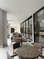 Un área de estar al aire libre con mesas y sillas, ventanas amplias y vistas al exterior.