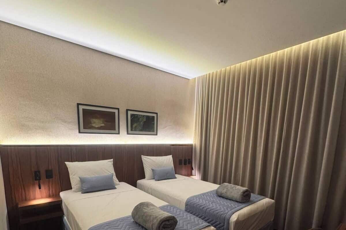 A hotel room with two beds, a wooden headboard, a ceiling light, and a window with curtains.