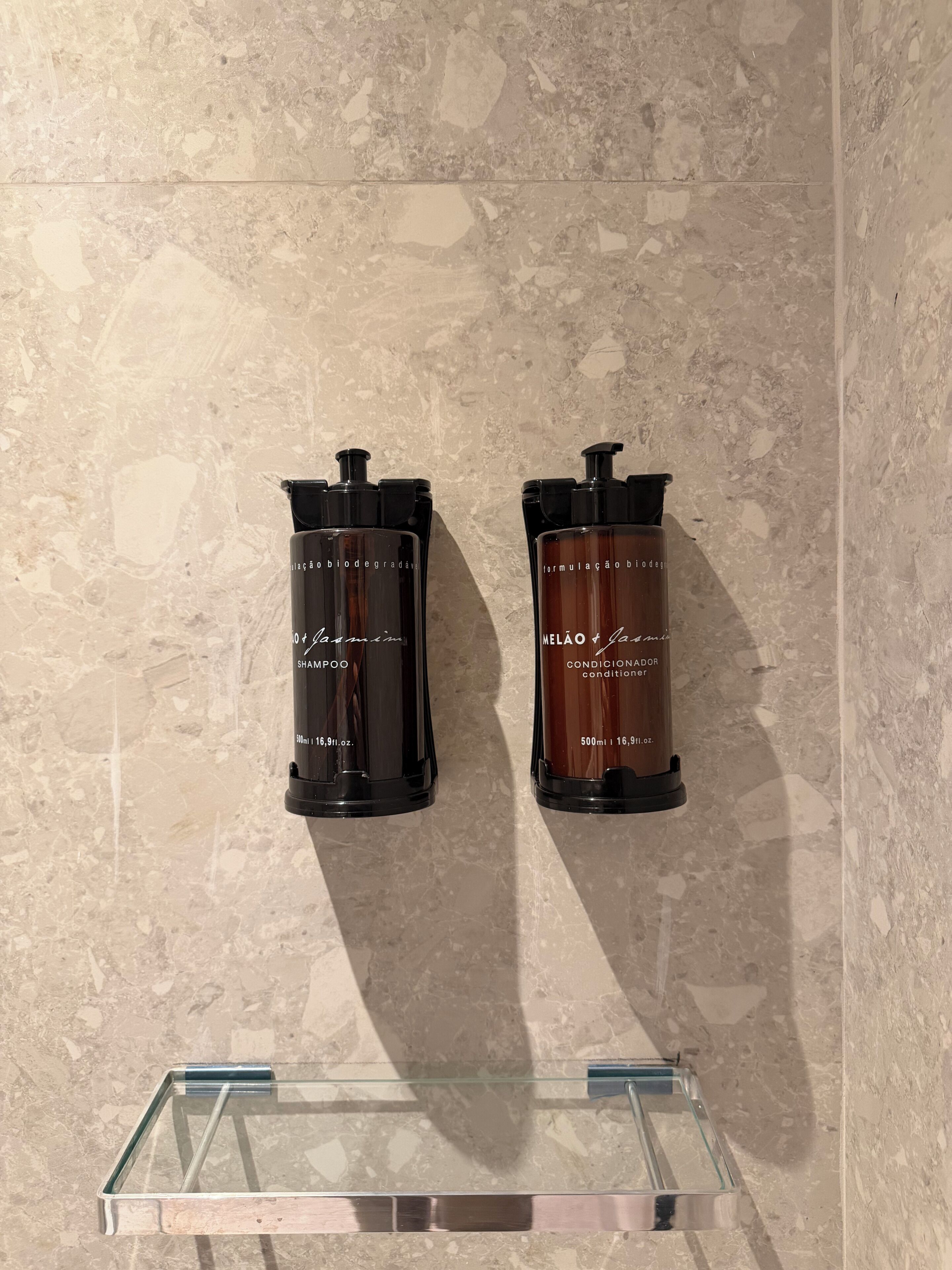 Dos botellas negras con etiquetas sobre una pared de mármol, una con la etiqueta 'SHAMPOO' y la otra 'CONDITIONER', con un estante de vidrio transparente debajo.