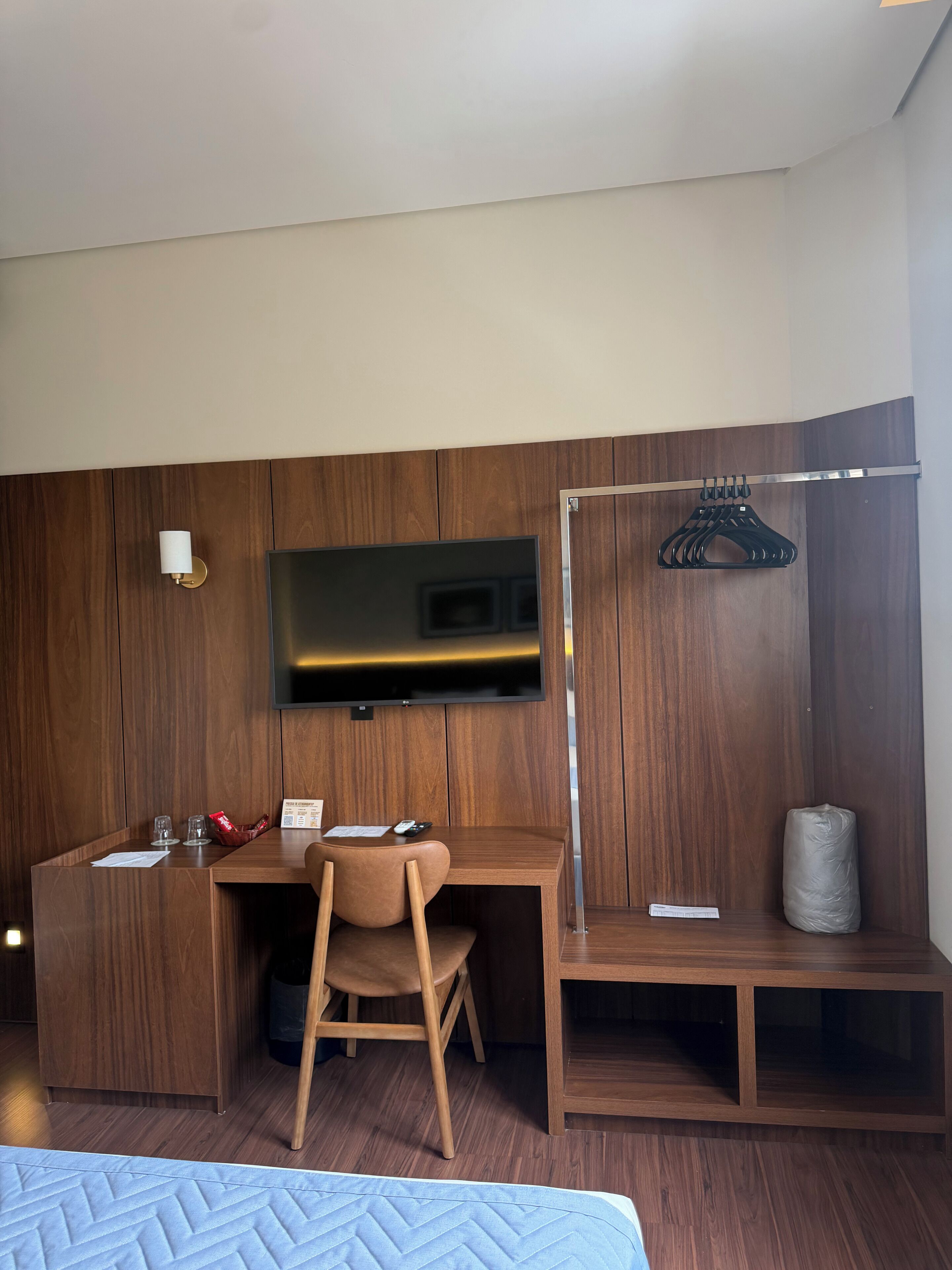 A hotel room with a wooden desk, a chair, a television mounted on the wall, and a clothing rack.