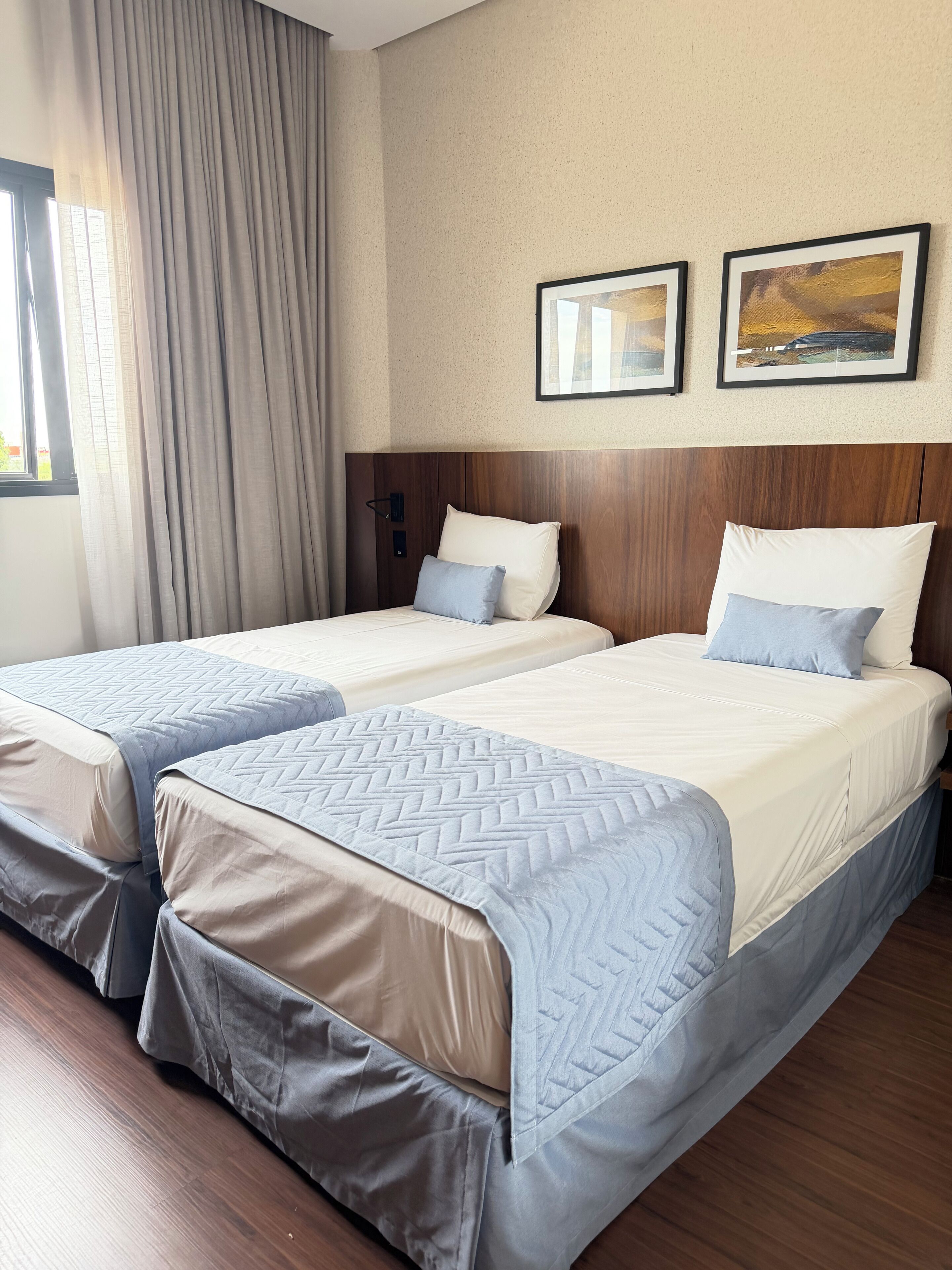 A hotel room with two beds, a wooden headboard, a window with curtains, and two framed pictures on the wall.