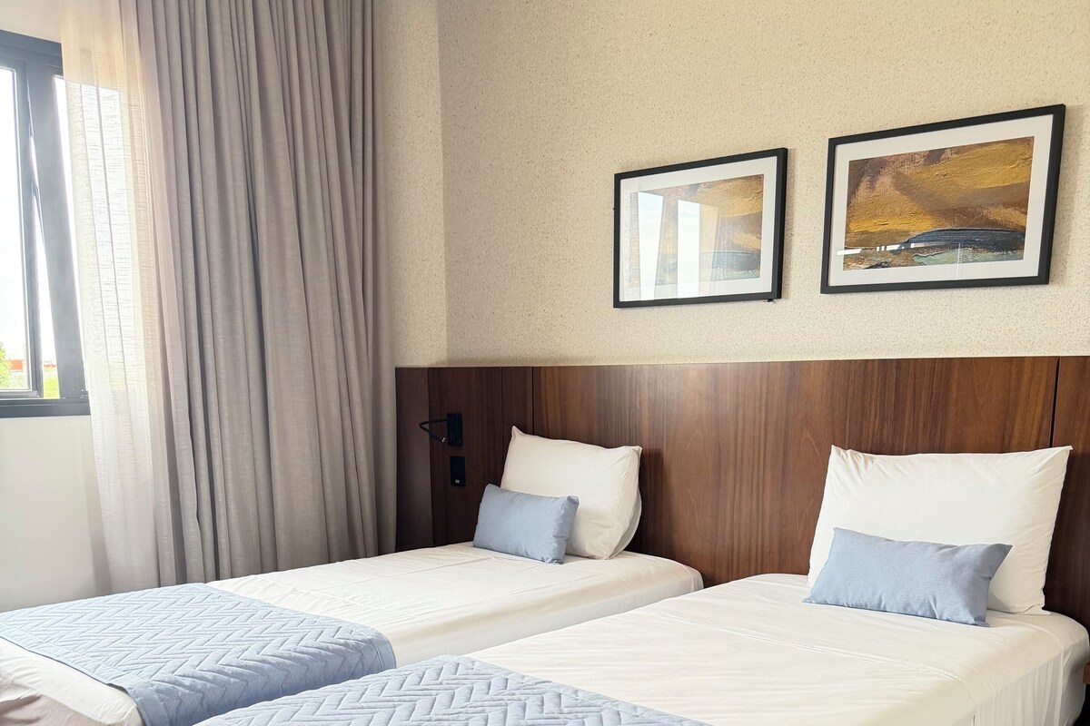 A hotel room with two beds, a wooden headboard, a window with curtains, and two framed pictures on the wall.