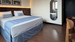 A hotel room with a bed, a desk, and a chair. There is a wall-mounted mirror and a door. - Class Hotel Americana (Americana)